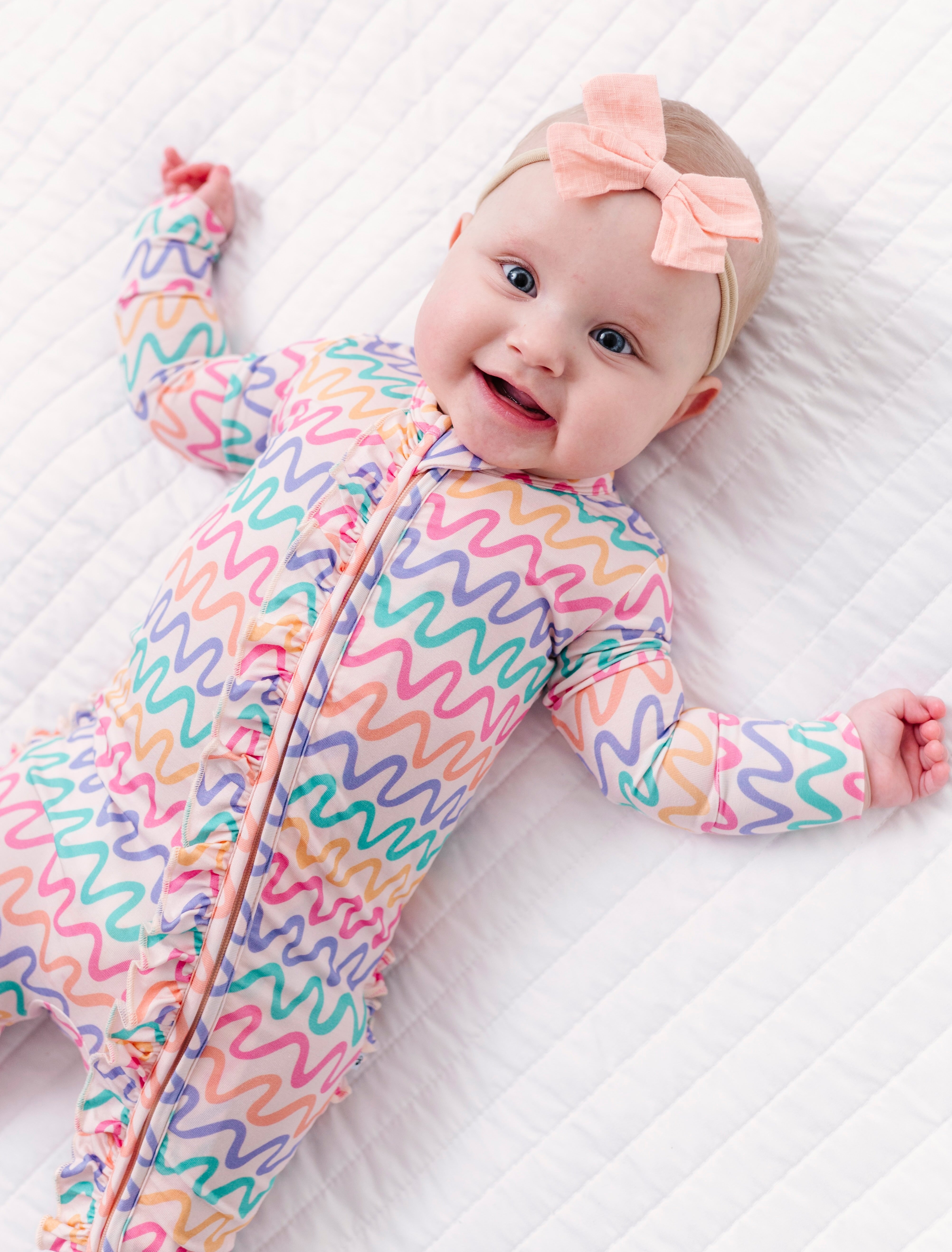 Baby wearing a colorful bamboo zip romper with a pink bow on a white quilted surface